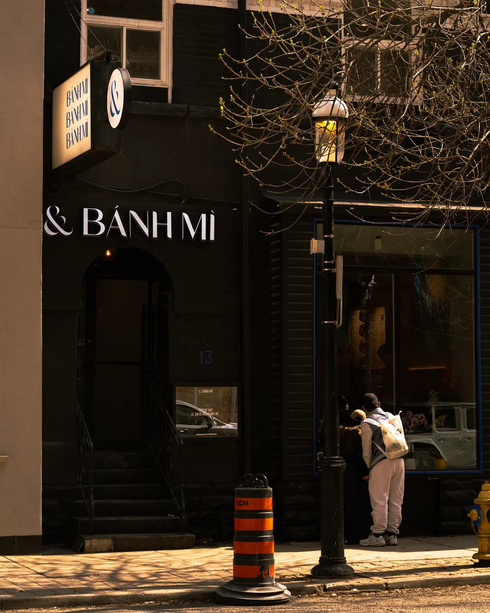 Exterior of And Banh Mi restaurant with a black facade, illuminated signage, steps leading to the entrance, a street lamp, and a person standing on the sidewalk.