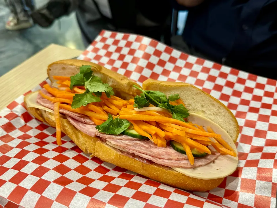 A Vietnamese banh mi sandwich filled with sliced cold cuts, pickled carrots, cucumber slices, and fresh cilantro sprigs, served on red and white checkered paper.