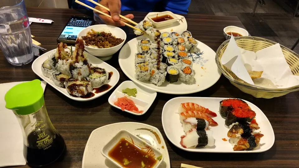 A wooden table filled with various Japanese dishes including sushi rolls, nigiri, a bowl of rice with meat, dipping sauces, and a bottle of soy sauce.