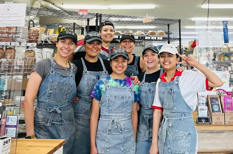 Six smiling female staff members wearing matching denim overalls and caps stand together behind the counter at The Bread Essentials bakery.