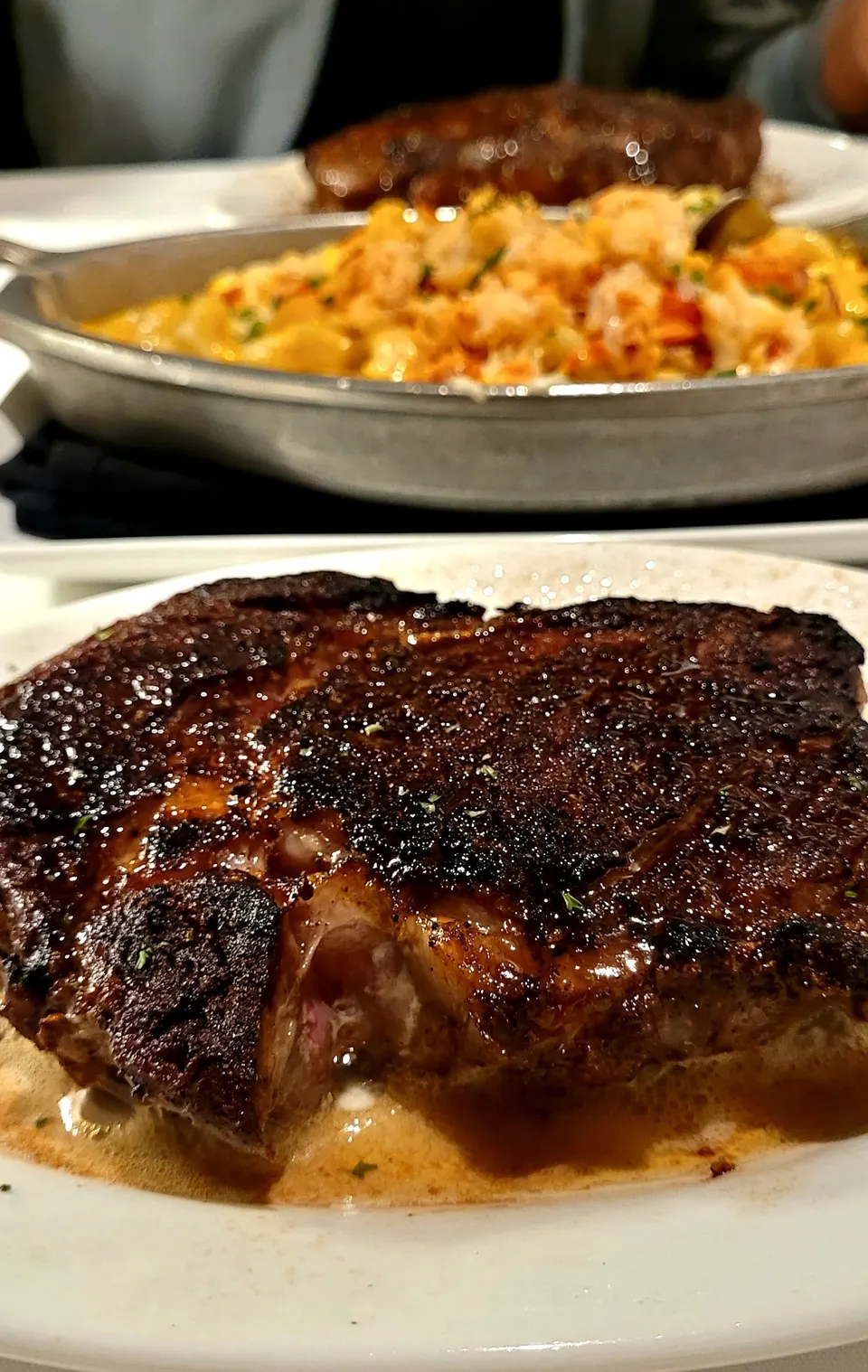 A thick, heavily seared ribeye steak served on a hot plate with sizzling butter, with a side dish of lobster mac and cheese in a metal pan blurred in the background.