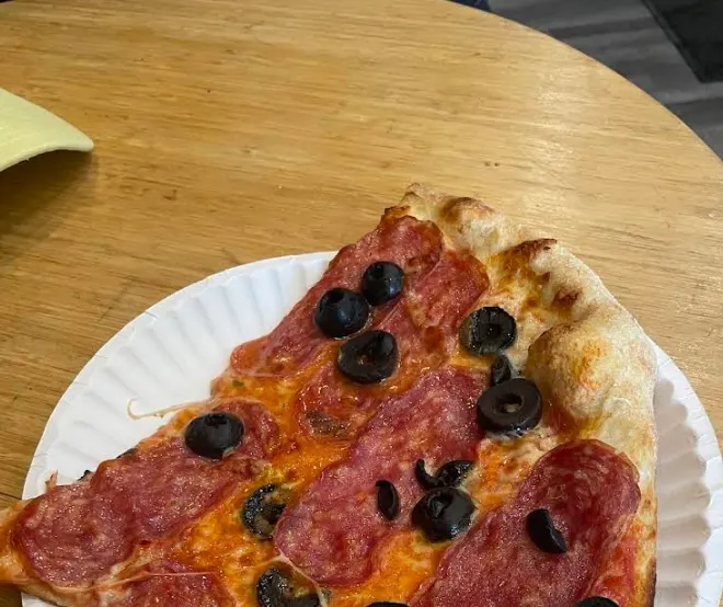 A slice of pepperoni and black olive pizza served on a white paper plate, resting on a light-colored wooden table.
