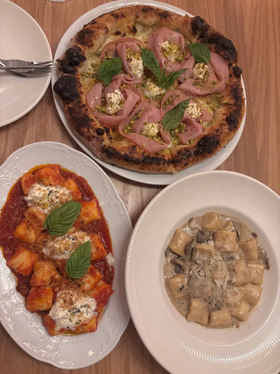 A top-down view of a wooden table featuring a gourmet pizza topped with mortadella, burrata, and pistachios, alongside two plates of gnocchi—one in a red tomato sauce with fresh basil and cheese, and another in a creamy mushroom sauce.