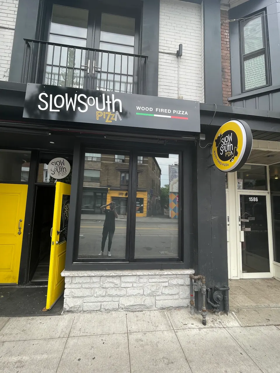 Exterior storefront of SlowSouth Pizza featuring a black facade, yellow doors, a stone base, and a round yellow sign hanging from the wall.