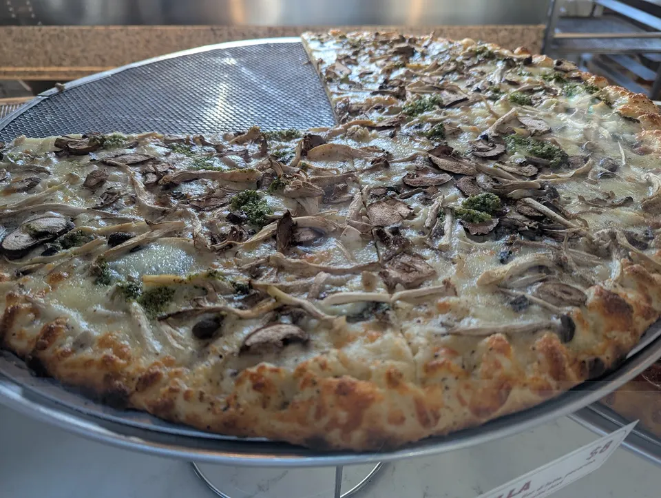 A large, thin-crust pizza topped with mushrooms, cheese, and dollops of pesto, with a few slices missing, sitting on a metal serving tray.