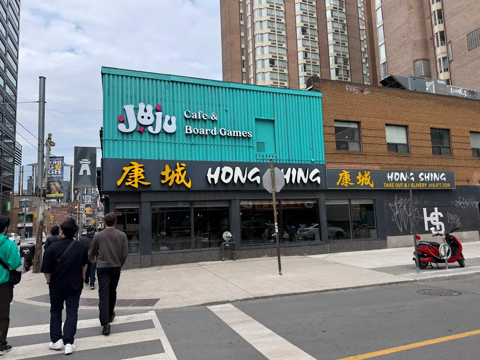 Street view of Hong Shing restaurant with a black facade and yellow Chinese characters, located below a teal-colored building for Juju Cafe & Board Games.