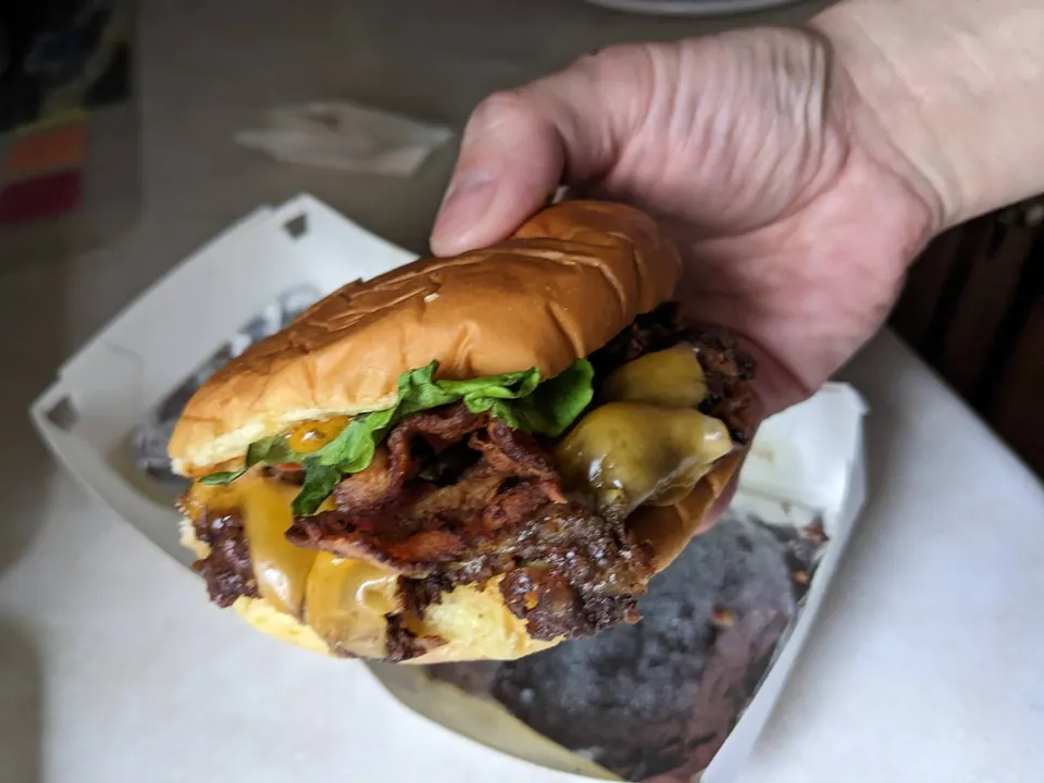 A hand holding a double cheeseburger with crispy bacon, melted cheese, and lettuce in a brioche bun, over a white takeout box.