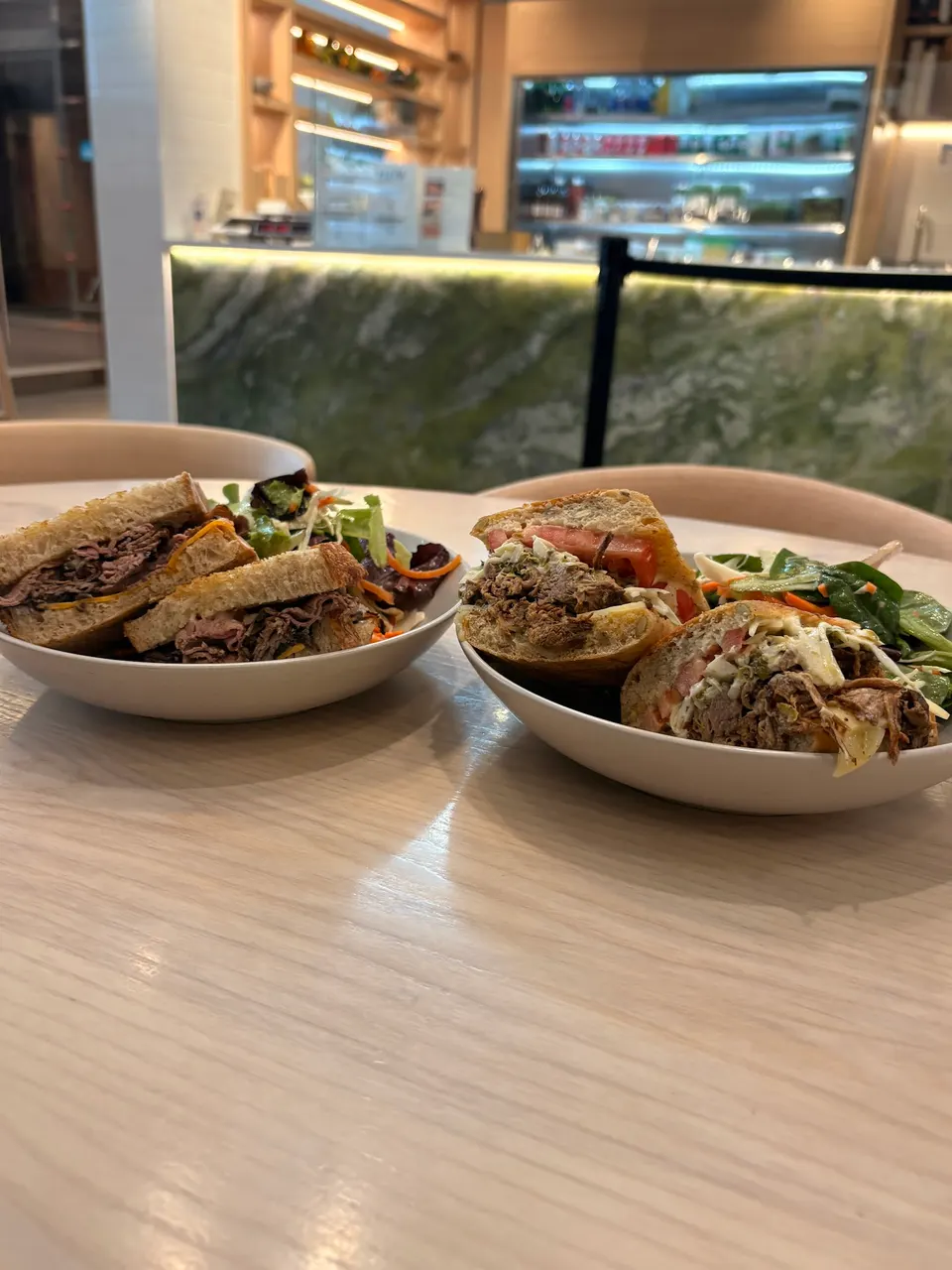 Two white bowls on a light wooden table, each containing a halved sandwich and a side salad, with a modern green marble counter in the background.