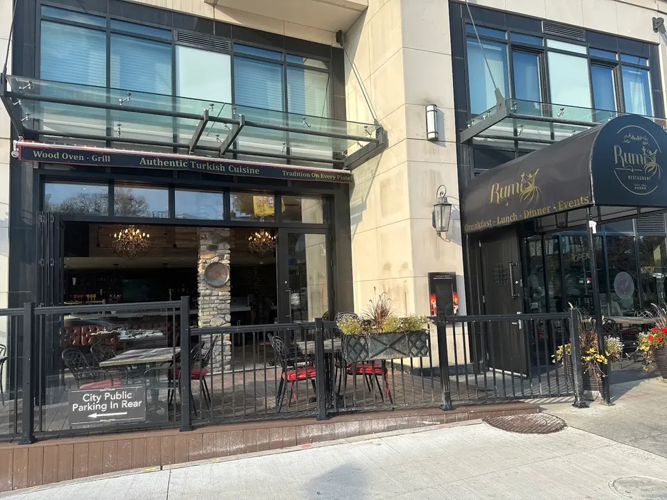 Exterior storefront of Rumi Restaurant featuring an outdoor patio with black metal tables and chairs, a dark awning with gold lettering, and large open glass doors leading inside.