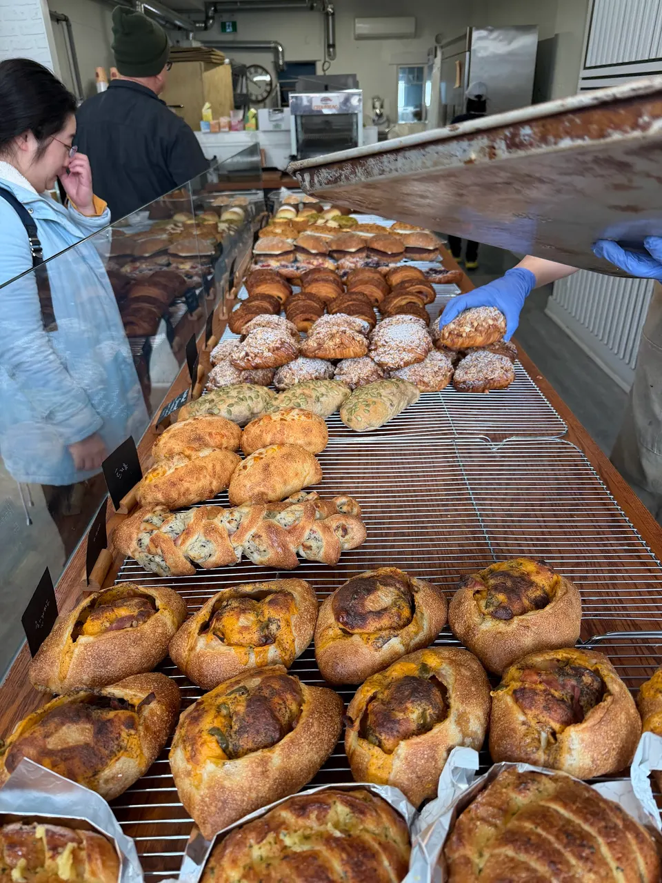 A wide selection of freshly baked artisanal breads, croissants, and pastries arranged on wire cooling racks on a wooden counter at Bricolage Bakery, with customers and staff in the background.