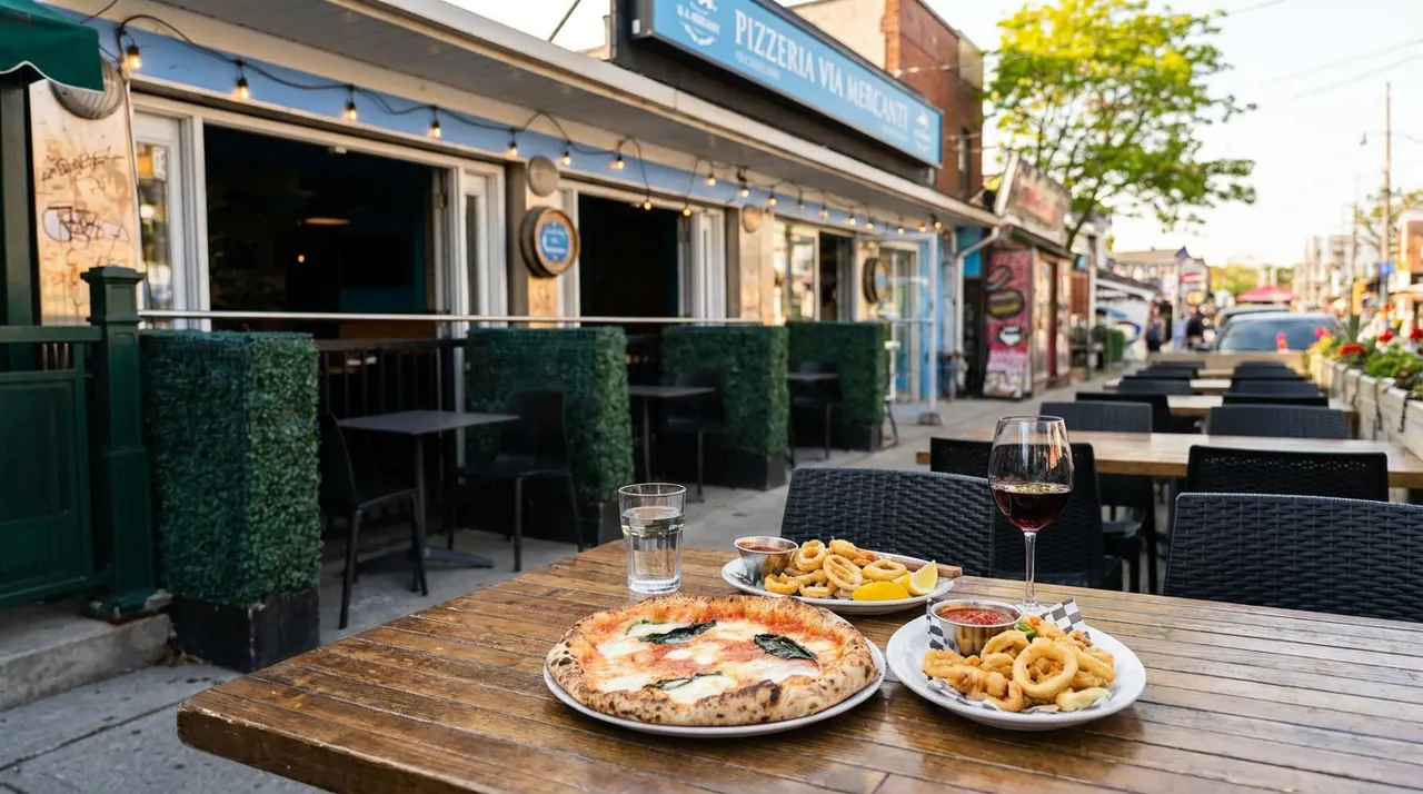 A wooden patio table set with a fresh Margherita pizza, two plates of fried calamari with dipping sauce, a glass of red wine, and a glass of water, in front of the Pizzeria Via Mercanti storefront.