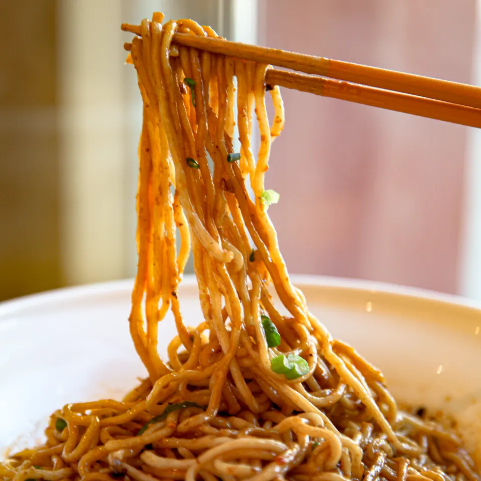 A close-up shot of noodles being lifted from a white bowl with wooden chopsticks, showing the texture and sauce coating the noodles.