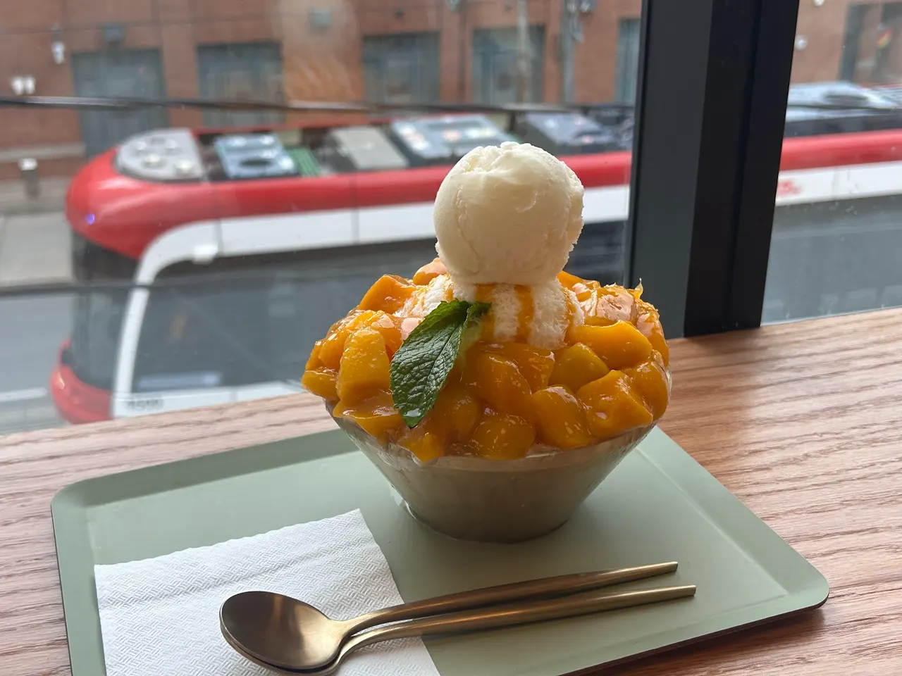 A bowl of mango bingsu topped with a scoop of vanilla ice cream and a mint leaf, served on a green tray with a spoon and napkin next to a window overlooking a city street.