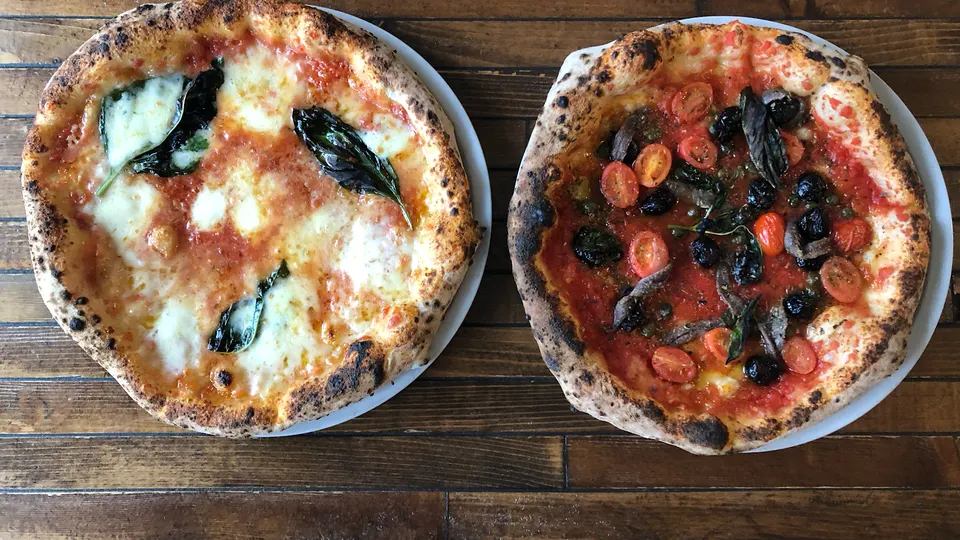 Two freshly baked Neapolitan pizzas served on white plates on a dark wooden table. The pizza on the left is a classic Margherita with melted mozzarella and fresh basil leaves. The pizza on the right is topped with cherry tomatoes, black olives, and anchovies.