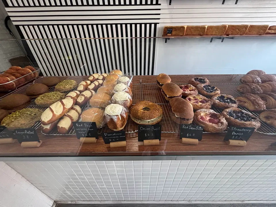 A bakery display case filled with various freshly baked pastries, including pretzels, cream-filled buns, onion basil bread, and fruit danishes, each labeled with small black price tags.