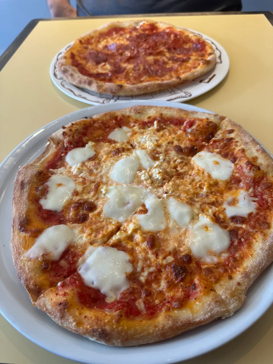 Two freshly baked pizzas served on white plates on a yellow table at Frank's Pizza House. The foreground pizza features melted mozzarella dollops on a tomato base, while the background pizza has pepperoni.