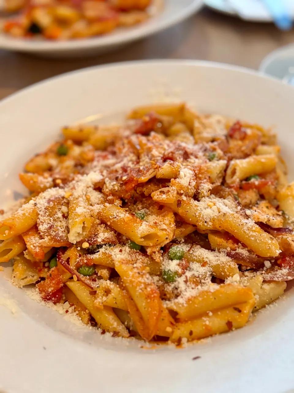 A close-up shot of a white bowl filled with penne pasta tossed in a tomato-based sauce, sprinkled with grated parmesan cheese, red pepper flakes, and green peas.