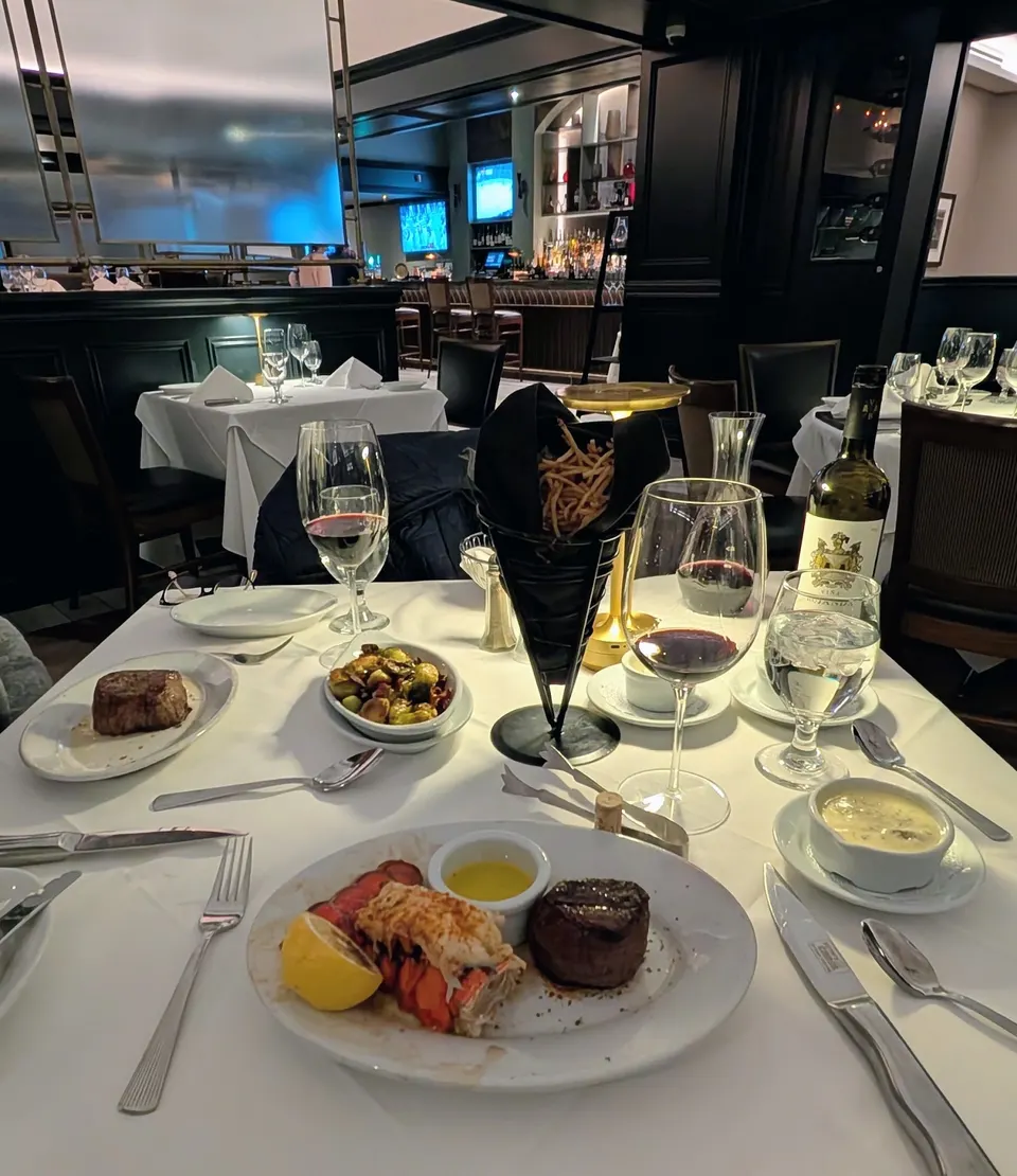 A white tablecloth table set for two at Ruth's Chris Steak House, featuring steak, lobster tail, brussels sprouts, french fries, red wine, and a bottle of wine.