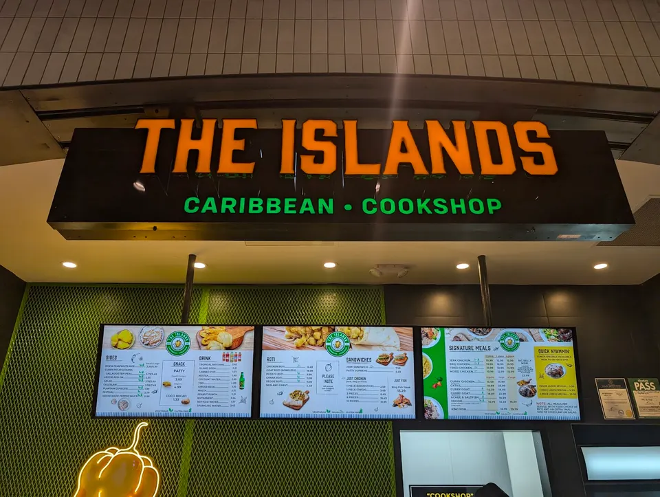 A brightly lit sign for 'THE ISLANDS CARIBBEAN COOKSHOP' hangs above three digital menu boards displaying various food and drink options. The menu boards show categories like 'Bites', 'Snack', 'Drink', 'Roti', 'Sandwiches', 'Signature Meals', and 'Quick Wraps'. The background features a green textur