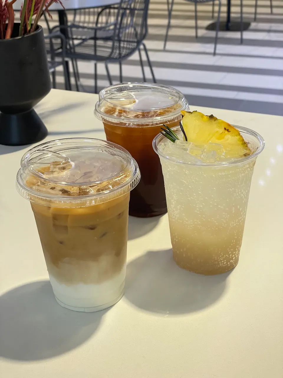 Three iced drinks in clear plastic cups on a white table: an iced latte with distinct milk and coffee layers, an iced black coffee, and a sparkling pineapple beverage garnished with a fresh pineapple slice and rosemary sprig.