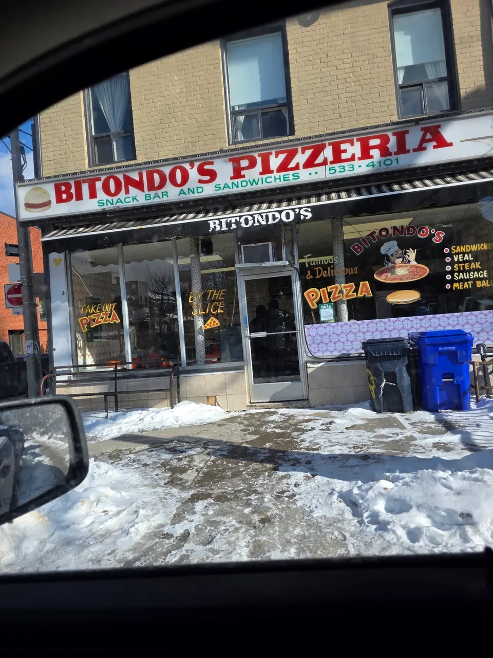 Exterior storefront of Bitondo's Pizzeria with a red and white sign, window decals advertising pizza by the slice, and snow on the ground in front.