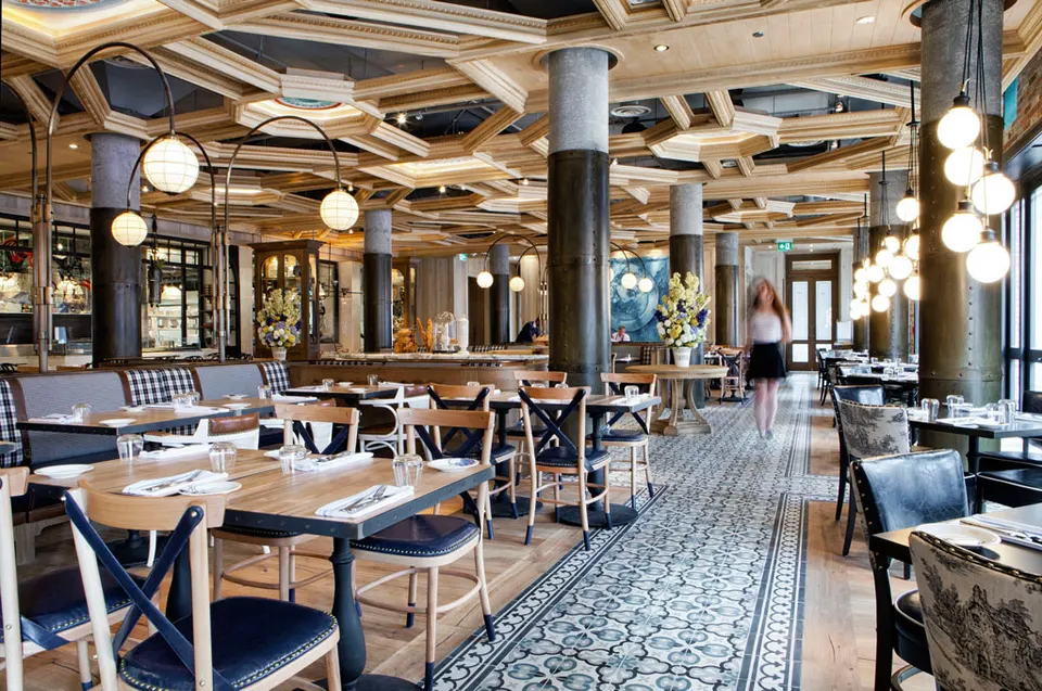 Spacious dining room at Cluny Bistro & Boulangerie featuring intricate wooden ceiling panels, patterned tile walkways, round columns, and elegant hanging globe lights over wooden tables.