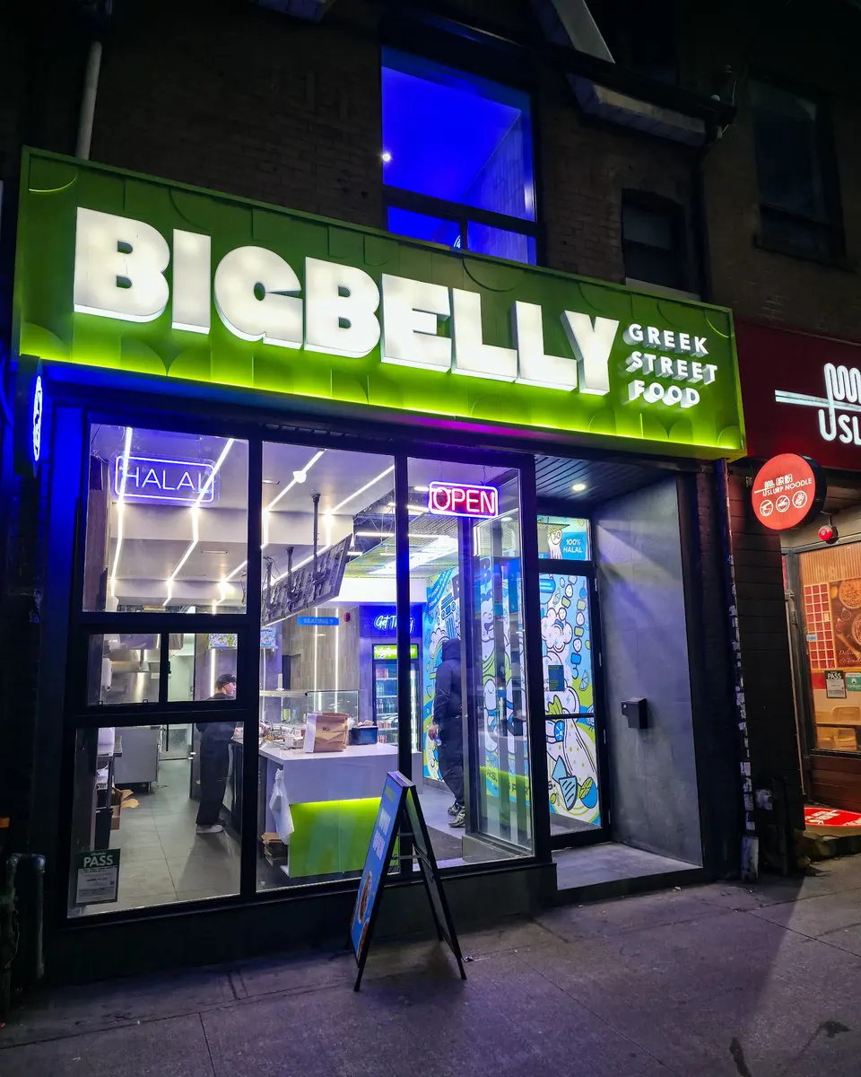 Brightly lit storefront of BIGBELLY Greek Street Food at night, featuring a vibrant green sign with white lettering, large glass windows showing the interior counter, and a sidewalk sandwich board sign.