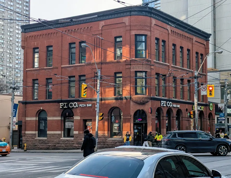 A three-story red brick building on a city corner featuring a Pi Co. Pizza Bar on the ground floor, with street traffic and pedestrians in the foreground.