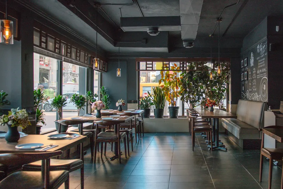 Interior view of Hong Shing Restaurant showing a modern dining area with dark walls, wooden tables set with plates, leather chairs, hanging pendant lights, and large potted plants near the windows.