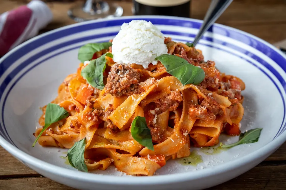 A bowl of wide ribbon pasta tossed in a rich meat sauce, topped with a scoop of white ricotta cheese and fresh green basil leaves.