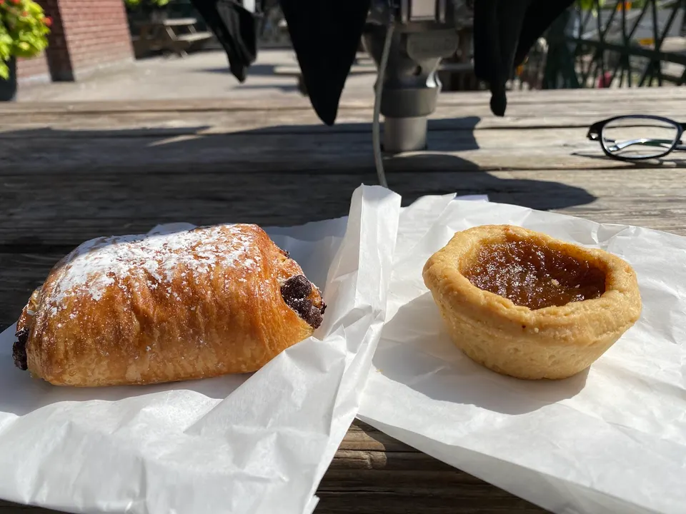 A pain au chocolat dusted with powdered sugar and a small jam tart, both resting on white paper sheets on a rustic wooden outdoor table.