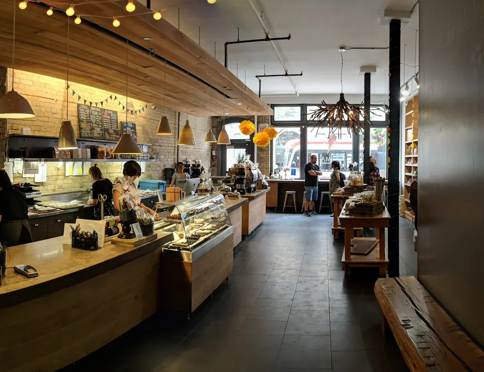 Interior of a modern chocolate shop with warm wood counters, hanging pendant lights, a glass display case filled with chocolates, and customers browsing in the background.