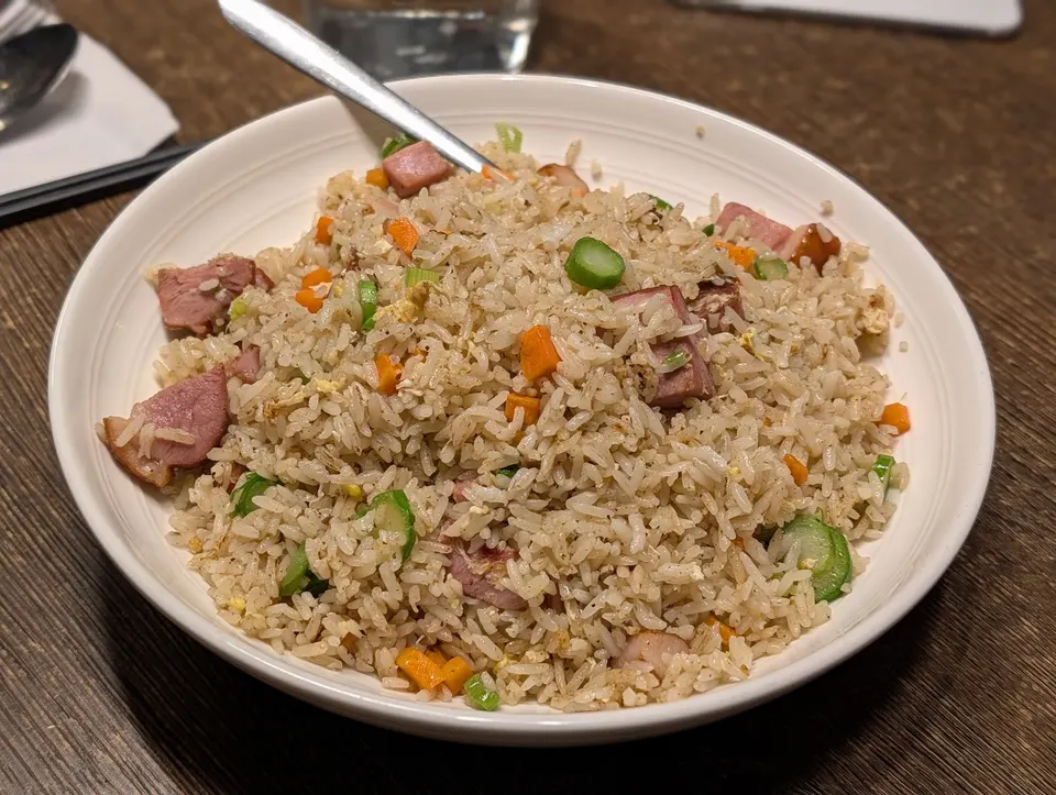 A white bowl filled with fried rice containing pieces of pork, chopped green onions, and diced carrots, served with a metal spoon on a wooden table.