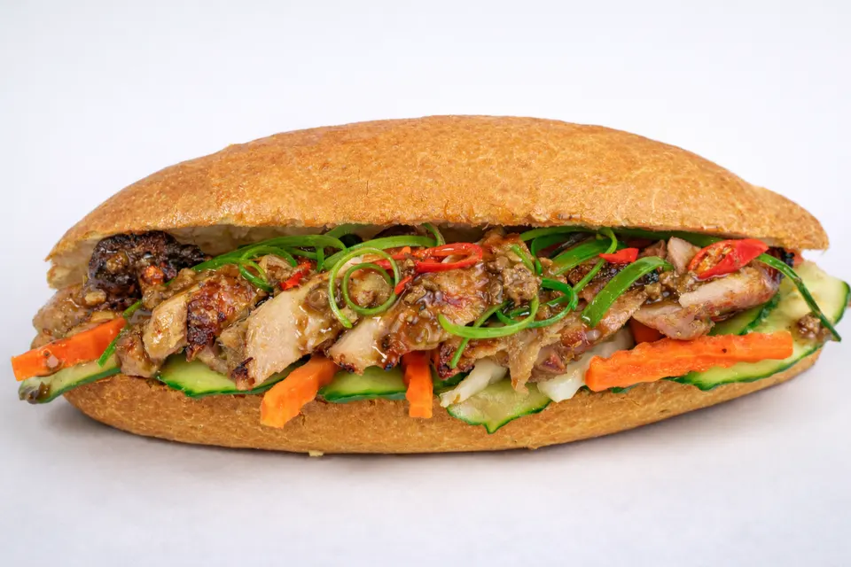 A close-up shot of a Vietnamese banh mi sandwich filled with grilled meat, pickled carrots, cucumber slices, green onions, and chili peppers on a white background.