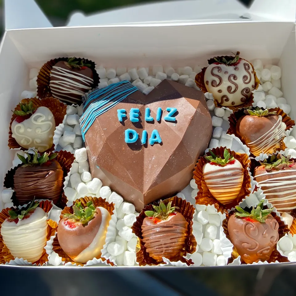 A white gift box filled with mini marshmallows, a large geometric chocolate heart with 'FELIZ DIA' written in blue, and assorted chocolate-covered strawberries decorated with white and dark chocolate drizzles.