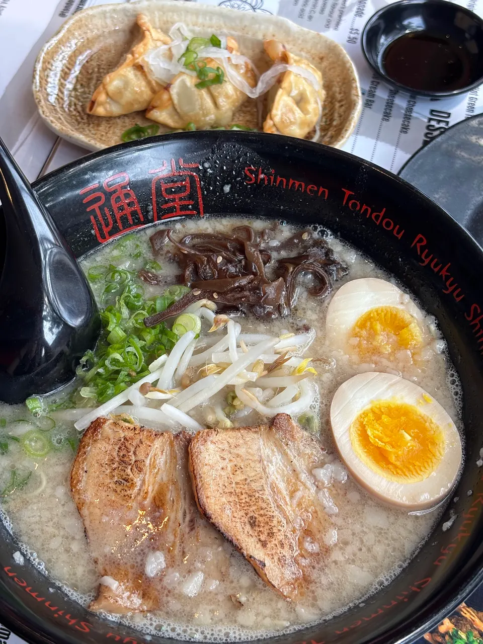 A bowl of tonkotsu ramen with chashu pork, soft-boiled egg halves, bean sprouts, green onions, and wood ear mushrooms, served alongside gyoza dumplings.