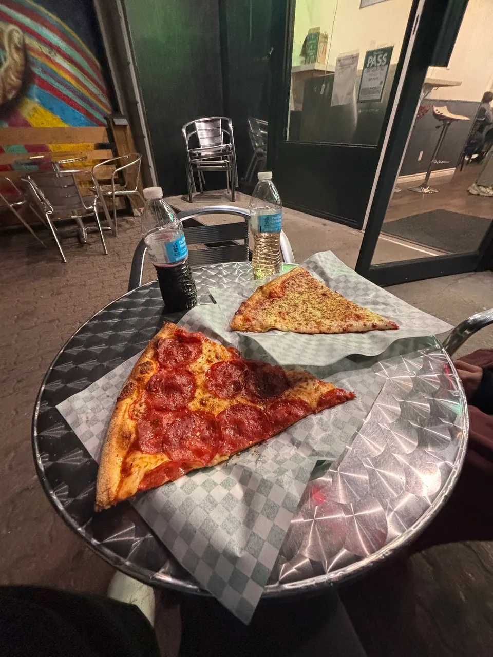 Two large slices of pizza, one pepperoni and one cheese, served on checkered paper on a round metal outdoor table next to two plastic water bottles.