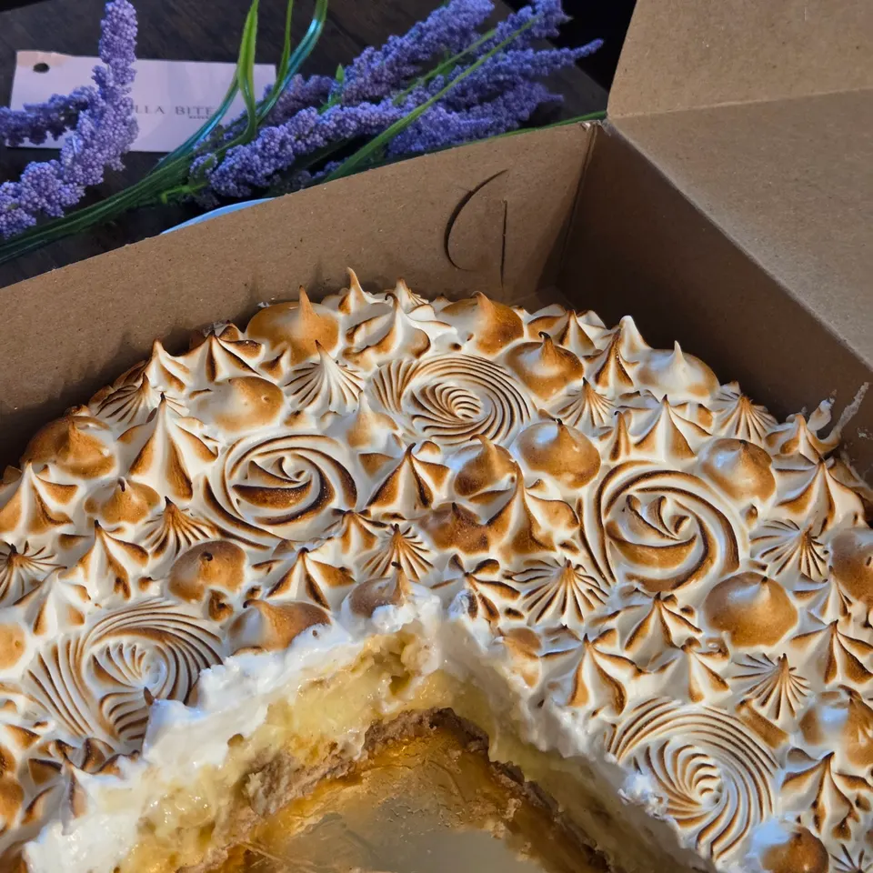 A close-up shot of a lemon meringue pie inside a cardboard bakery box, with a slice already removed to show the yellow filling and crust. Lavender flowers are visible in the background.