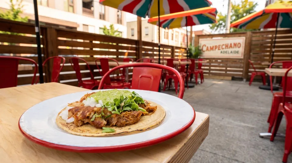A close-up of a single taco topped with meat, onions, cilantro, and green salsa on a white plate with a red rim, set on a wooden table at an outdoor patio with red chairs and colorful umbrellas.