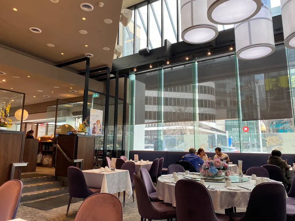 Spacious dining room at Moon Palace Cantonese Cuisine featuring high ceilings, large windows looking out to the city, round tables with white tablecloths, and purple chairs.