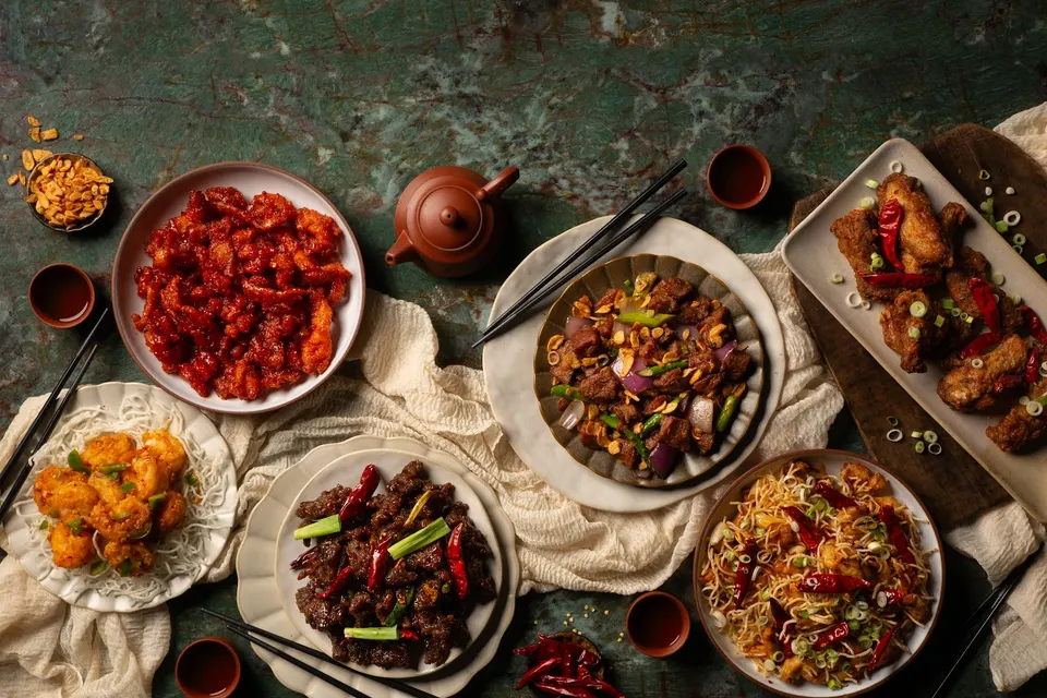 An overhead view of a Chinese feast featuring sweet and sour chicken, black pepper beef, crispy noodles, fried chicken wings, and stir-fried shrimp on a green marble table with a teapot.