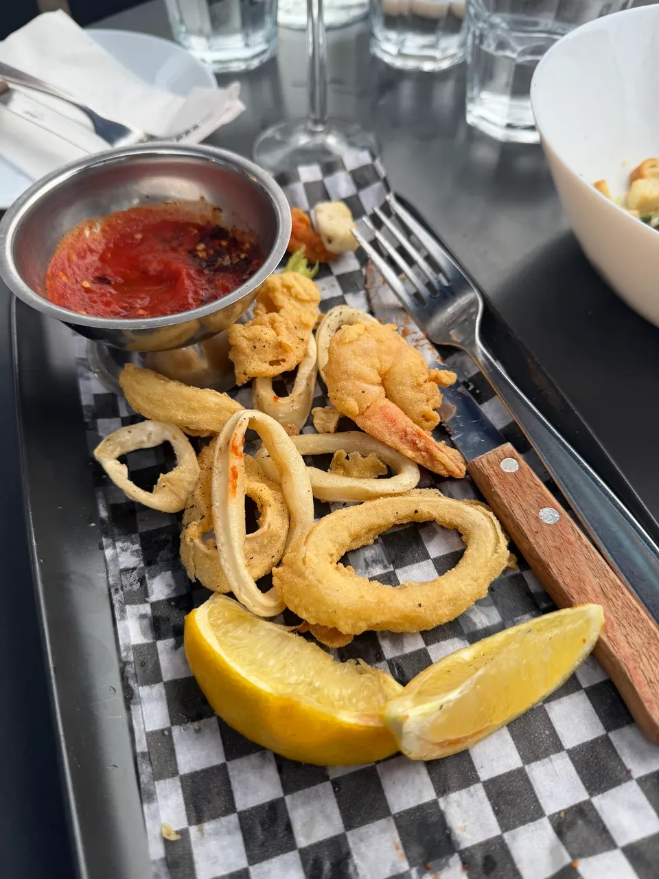 A platter of crispy fried calamari rings and a single fried shrimp served with lemon wedges, a small metal bowl of red dipping sauce, and cutlery on a black and white checkered paper liner.