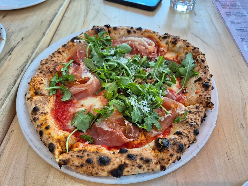 A freshly baked Neapolitan-style pizza on a white plate, topped with tomato sauce, melted mozzarella, prosciutto slices, fresh arugula, and grated cheese.