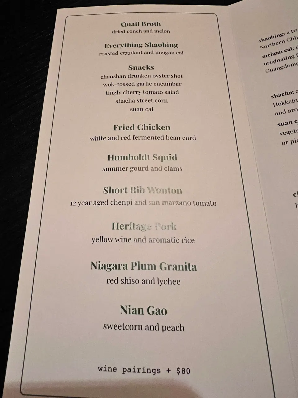 A close-up shot of a printed tasting menu from Yan Dining Room, featuring courses like Quail Broth, Everything Shaobing, Snacks, Fried Chicken, Humboldt Squid, Short Rib Wonton, Heritage Pork, Niagara Plum Granita, and Nian Gao.