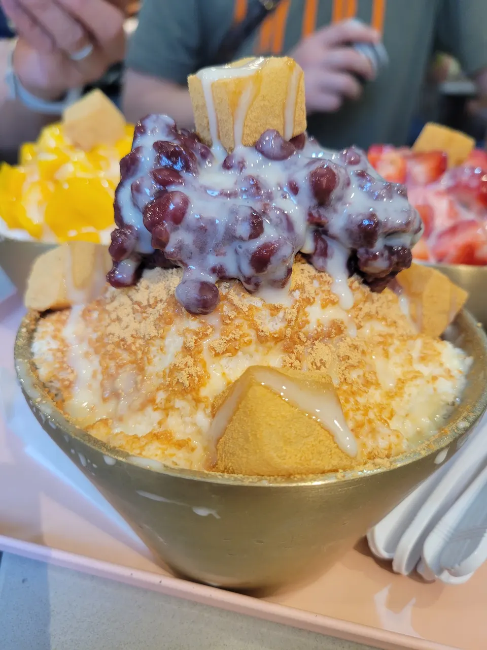 A close-up of a large bowl of Korean bingsu shaved ice topped with red beans, condensed milk, injeolmi powder, and rice cake cubes, served in a brass bowl.