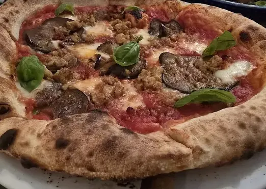 A close-up view of a freshly baked Neapolitan-style pizza with a thick, charred crust, topped with tomato sauce, melted mozzarella, savory sausage, sliced eggplant, and fresh basil leaves.