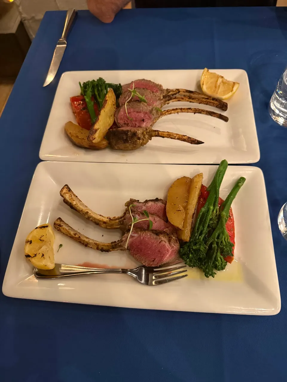Two white rectangular plates of grilled lamb chops served with roasted potatoes, broccolini, and a lemon wedge on a blue tablecloth.