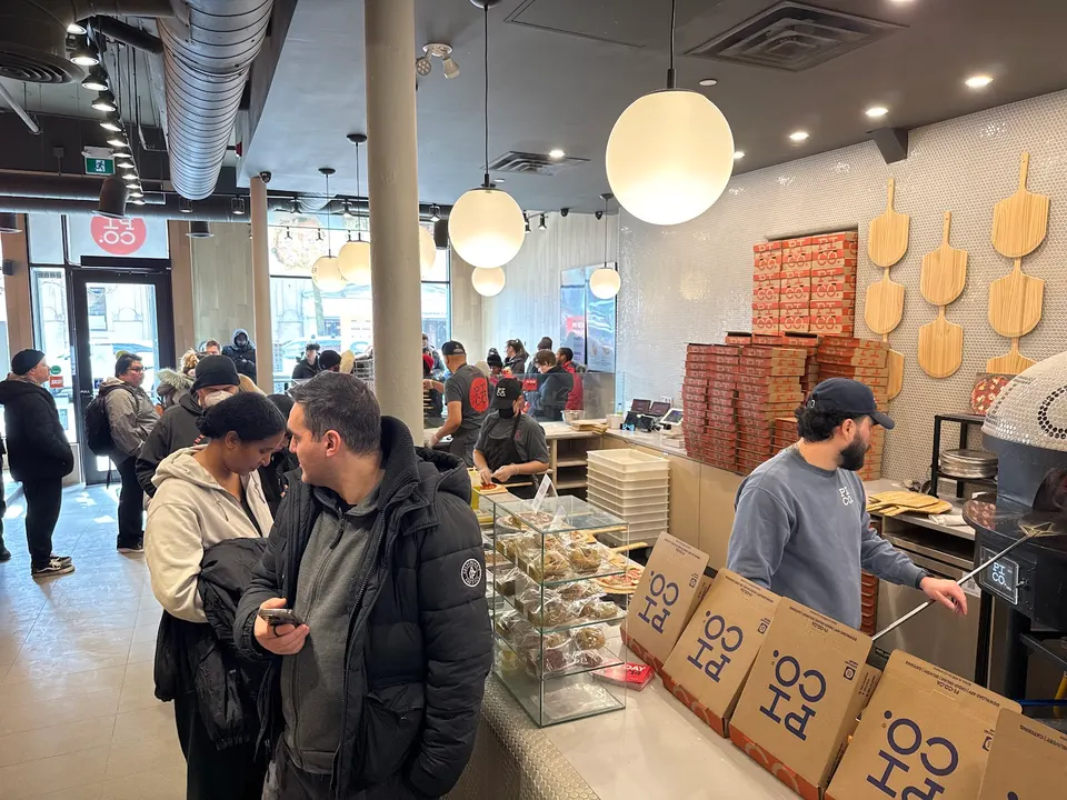 Customers waiting in line inside a busy Pi Co. Pizza Bar location, with staff preparing pizzas behind the counter and stacks of pizza boxes nearby.