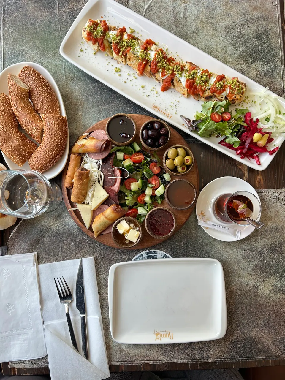 An overhead view of a Turkish breakfast and meze spread at Rumi Restaurant, featuring a long plate of beyti kebab, a round wooden platter with cheese, olives, and salad, a plate of simit bread, and Turkish tea.