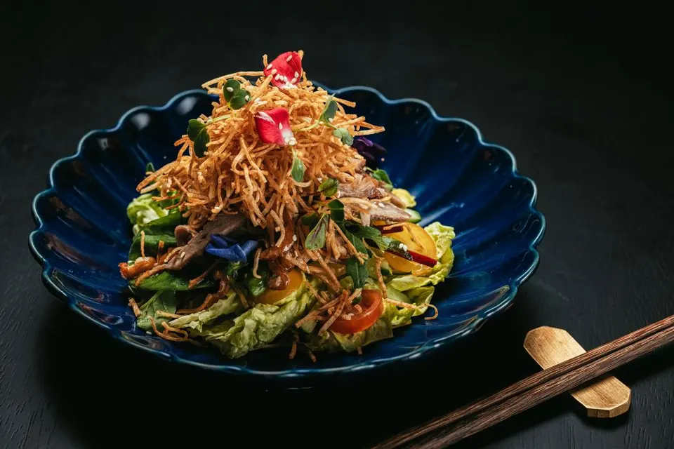 A gourmet salad topped with crispy fried potato shreds, fresh greens, cherry tomatoes, and edible flower petals, served in a scalloped dark blue bowl next to wooden chopsticks.